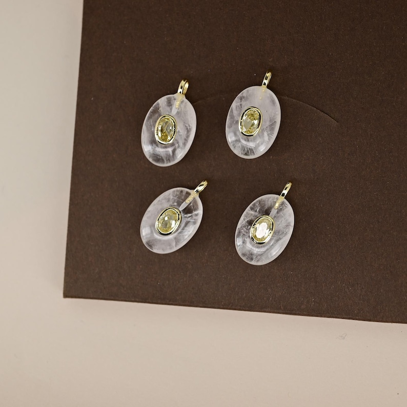 May include: Four oval-shaped pendants with a gold bail and a central yellow gemstone. The pendants are set in a clear, slightly cloudy material, possibly quartz or resin. The background is a dark brown.