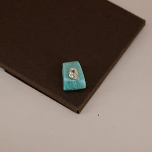 Pu&ograve; includere: Un ciondolo trapezoidale in pietra turchese con una gemma trasparente di forma ovale al centro, incastonata in una cornice di metallo dorato. Il ciondolo presenta una superficie liscia e lucida con una colorazione blu-verde e venature.