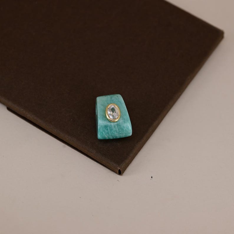 Pu&ograve; includere: Un ciondolo trapezoidale in pietra turchese con una gemma trasparente di forma ovale al centro, incastonata in una cornice di metallo dorato. Il ciondolo presenta una superficie liscia e lucida con una colorazione blu-verde e venature.