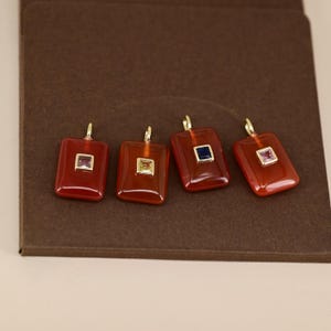 Op de afbeelding: Vier rechthoekige hangers met een roodbruine tint, elk met een kleine, vierkante edelsteen in verschillende kleuren: rood, geel, blauw en roze. De hangers hebben goudkleurige lussen aan de bovenkant om op te hangen.