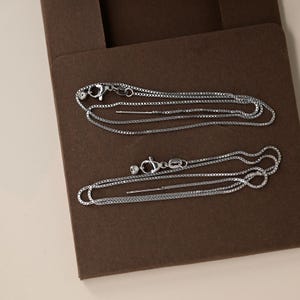 Puede incluir: Dos collares plateados con cadenas tipo caja, cierres de mosquetón y pequeñas cuentas redondas. Los collares se exhiben sobre una superficie rectangular marrón, posiblemente dentro de una caja de joyería.