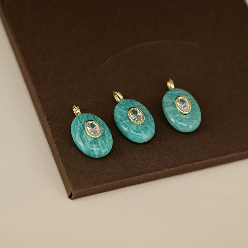 May include: Three oval-shaped pendants with a turquoise stone and a central clear stone set in a gold-colored metal. The pendants are displayed on a brown surface, showcasing their design and color.