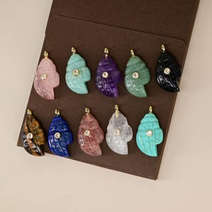 Op de afbeelding: Een verzameling schelpvormige hangers in verschillende kleuren, waaronder roze, turquoise, paars en zwart. Elke hanger heeft een kleine, ronde, heldere steen en een goudkleurige beugel. De hangers worden gepresenteerd op een bruin oppervlak.