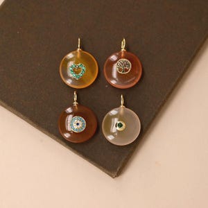May include: Four circular pendants featuring smooth, translucent stones in shades of amber, reddish-brown, dark brown, and pale gray. Each pendant has a gold-toned bail and a central gold-toned charm: a heart with green stones, a tree, an eye, and a green gem.