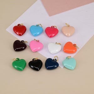 Puede incluir: Una colección de colgantes en forma de corazón en varios colores, incluyendo rojo, azul, rosa y verde. Cada colgante tiene un lazo dorado para sujetarlo a joyas u otras manualidades. Los colgantes son brillantes y parecen estar hechos de resina o esmalte.