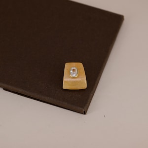Pu&ograve; includere: Un anello di forma trapezoidale con una base in pietra arancione liscia e traslucida. Al centro della pietra arancione si trova una piccola gemma trasparente di forma ovale incastonata in una sottile montatura in metallo dorato lucido.