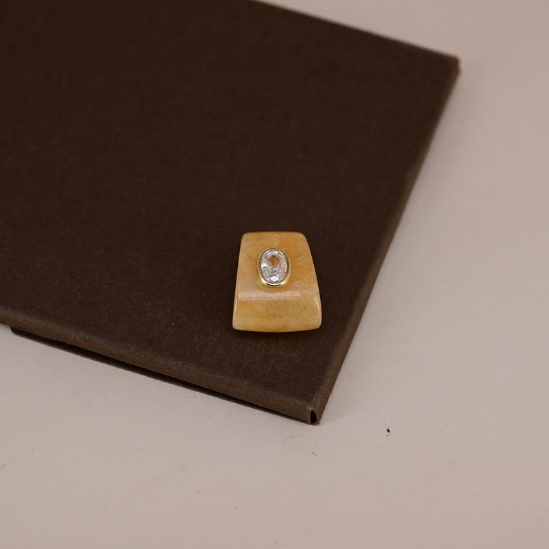 Pu&ograve; includere: Un anello di forma trapezoidale con una base in pietra arancione liscia e traslucida. Al centro della pietra arancione si trova una piccola gemma trasparente di forma ovale incastonata in una sottile montatura in metallo dorato lucido.