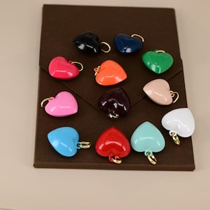 May include: A collection of heart-shaped charms in various colors, including red, pink, blue, green, and black. Each charm has a gold-colored loop at the top. The charms are arranged on a brown surface.