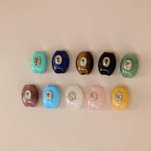 Puede incluir: Diez cuentas de forma ovalada dispuestas en dos filas. Cada cuenta presenta un engaste central en tono dorado que contiene un cristal transparente facetado. Las cuentas vienen en varios colores, incluyendo turquesa, azul oscuro, marrón, negro, verde, rojo, azul claro, blanco, rosa y amarillo.