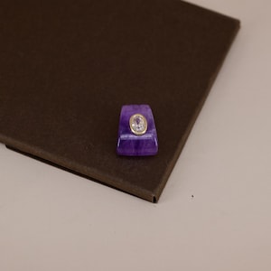 Pu&ograve; includere: Un ciondolo in pietra viola di forma trapezoidale con una superficie liscia e lucida caratterizzata da venature naturali visibili. Al centro della pietra viola si trova una piccola gemma trasparente ovale incastonata in una sottile montatura dorata.