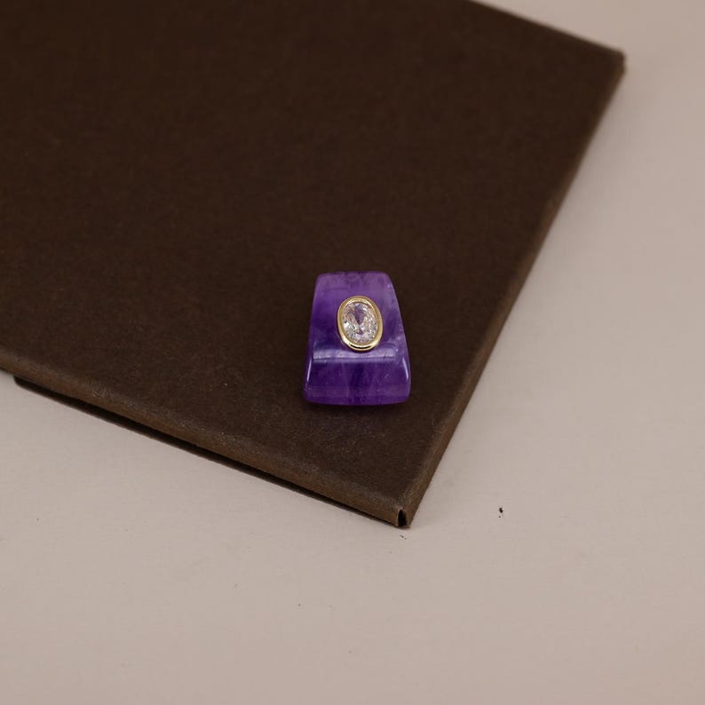 Pu&ograve; includere: Un ciondolo in pietra viola di forma trapezoidale con una superficie liscia e lucida caratterizzata da venature naturali visibili. Al centro della pietra viola si trova una piccola gemma trasparente ovale incastonata in una sottile montatura dorata.