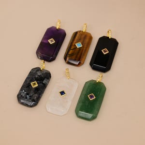 Pode incluir: Seis pingentes retangulares com pedras preciosas polidas em roxo, olho de tigre marrom, preto, cinza salpicado, transparente e verde. Cada pingente tem um suporte dourado com pequenas pedras claras e um detalhe central de pedra quadrada.