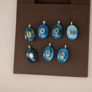 May include: Eight oval-shaped pendants with a gold bail. Each pendant features a blue stone with a smaller, lighter-colored stone in the center. The pendants are displayed on a brown surface.