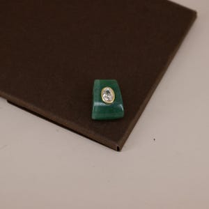 Pu&ograve; includere: Un componente per anello in pietra verde di forma trapezoidale con una gemma trasparente ovale incastonata in una montatura color oro. L'oggetto presenta una superficie liscia e lucida e una forma geometrica distinta, con una tonalit&agrave; verde.