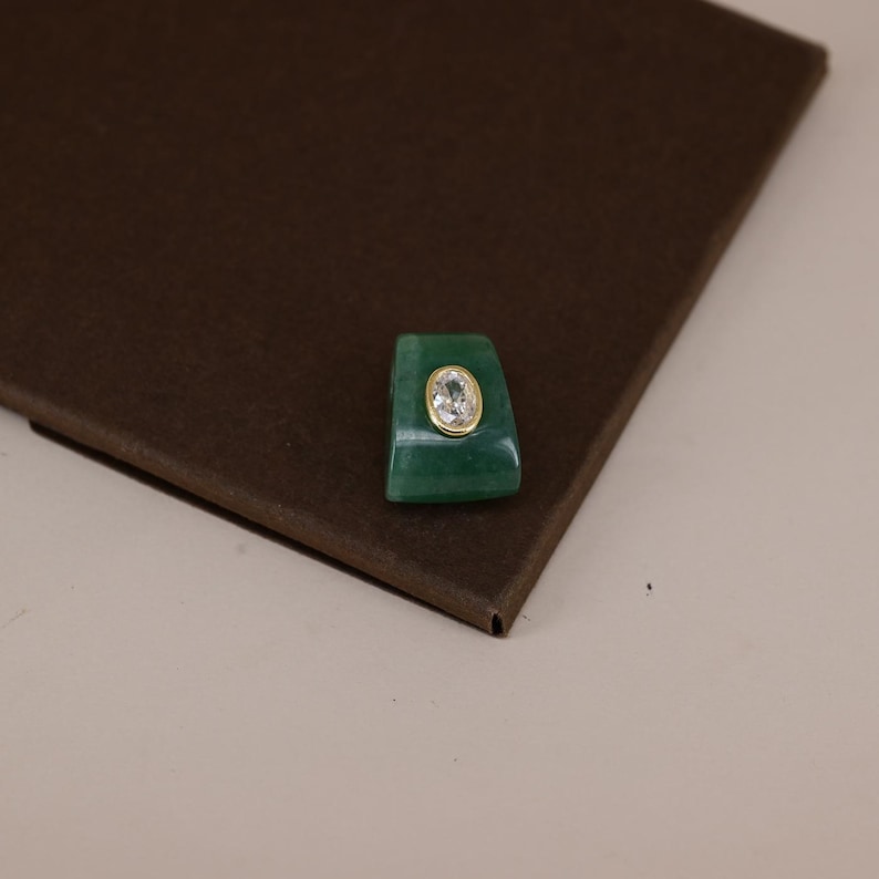 Pu&ograve; includere: Un componente per anello in pietra verde di forma trapezoidale con una gemma trasparente ovale incastonata in una montatura color oro. L'oggetto presenta una superficie liscia e lucida e una forma geometrica distinta, con una tonalit&agrave; verde.