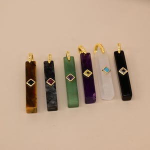Pode incluir: Seis pingentes retangulares em forma de barra em várias cores, incluindo marrom, cinza escuro, verde, roxo, transparente e preto. Cada pingente possui um gancho dourado com pequenas pedras claras e uma gema quadrada em moldura dourada.