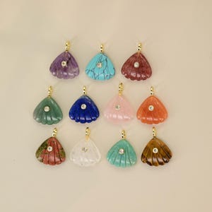 Op de afbeelding: Een verzameling edelsteenhangers in verschillende kleuren, waaronder paars, turquoise en rood. Elke hanger heeft een goudkleurige beugel en een kleine, ronde, heldere steen. De hangers zijn schelpvormig.