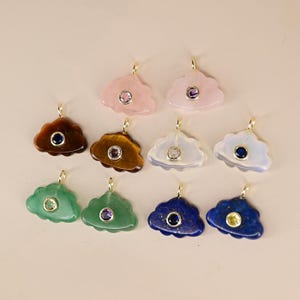 May include: A collection of cloud-shaped pendants with gold-colored bails. The pendants are made of various gemstones, including rose quartz, tiger's eye, and lapis lazuli, each with a central gemstone accent. The color palette includes pink, brown, green, and blue.