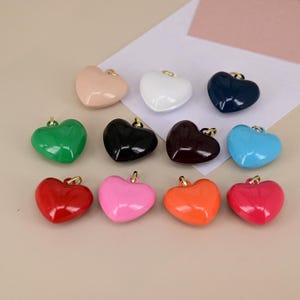 May include: A collection of heart-shaped charms in various colors, including pink, red, orange, green, blue, black, white, and brown. Each charm has a gold-colored loop at the top for attaching to jewelry or other crafts.