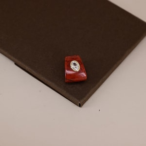 Pu&ograve; includere: Un ciondolo di forma trapezoidale caratterizzato da una pietra liscia rosso scuro con finitura lucida. Al centro della pietra rossa si trova una piccola gemma trasparente di forma ovale incastonata in una sottile cornice metallica dorata.