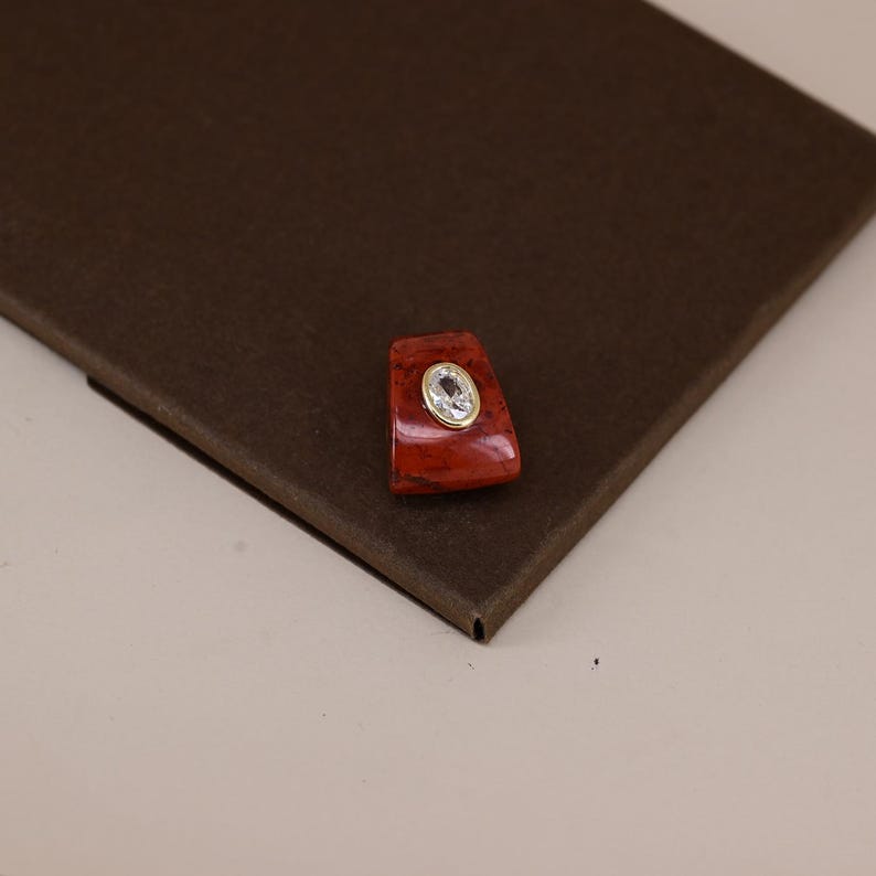 Pu&ograve; includere: Un ciondolo di forma trapezoidale caratterizzato da una pietra liscia rosso scuro con finitura lucida. Al centro della pietra rossa si trova una piccola gemma trasparente di forma ovale incastonata in una sottile cornice metallica dorata.