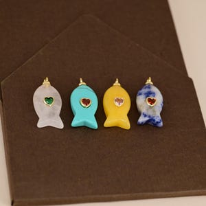 Puede incluir: Cuatro colgantes con forma de pez en piedra blanca, turquesa, amarilla y azul-gris. Cada colgante presenta una piedra preciosa en forma de corazón y detalles dorados. Los colgantes se muestran sobre una superficie marrón.