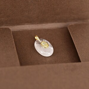 May include: A clear, oval-shaped pendant with a gold bail. The pendant features a smaller, oval-shaped yellow gemstone set in a gold bezel. The pendant is displayed on a brown surface.