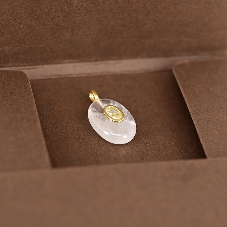 May include: A clear, oval-shaped pendant with a gold bail. The pendant features a smaller, oval-shaped yellow gemstone set in a gold bezel. The pendant is displayed on a brown surface.