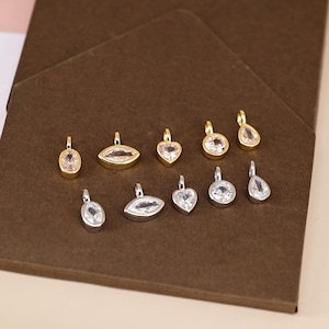 May include: A selection of small pendants featuring clear, faceted gemstones in diverse shapes, such as oval, marquise, heart, round, and teardrop. Some pendants have gold-coloured settings, while others are silver-coloured. The pendants are arranged on a brown surface.