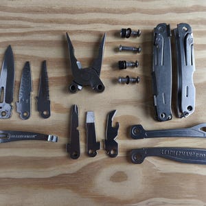 Könnte beinhalten: Eine Sammlung silberner Multitools und Zubehörteile, darunter Messer, Zangen und Schraubendreher, angeordnet auf einer hellbraunen Holzoberfläche. Die Werkzeuge sind in verschiedenen Größen und Formen erhältlich.