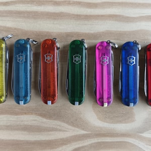 Könnte beinhalten: Sieben farbige Schweizer Taschenmesser in Gelb, Blau, Orange, Grün, Pink, Dunkelblau und Rot. Jedes Messer hat ein silbernes Emblem und einen kleinen Schlüsselring. Sie liegen auf einer hellbraunen Holzoberfläche.