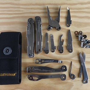 May include: A collection of silver multi-tool components, such as pliers, blades, and screwdrivers, displayed on a wooden surface. A black nylon sheath with the "LEATHERMAN" logo is also visible. The tools are made of metal.