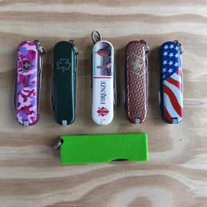 Pode incluir: Uma coleção de canivetes com vários designs, incluindo um padrão de camuflagem rosa, um trevo verde, uma vista panorâmica com a palavra "FIRENZE", um padrão vermelho e dourado e uma bandeira americana. Uma faca verde na parte inferior.