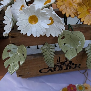 Puede incluir: Una caja de madera con la etiqueta Brasserie Caulier contiene flores de cerámica con pétalos blancos y amarillos, junto a flores amarillas. De la caja cuelgan hojas de cerámica verde tipo monstera y helecho con acabado brillante.