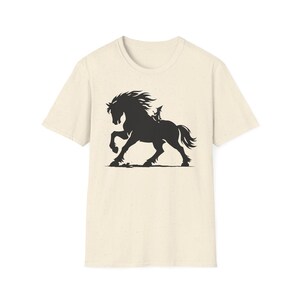 May include: Cream-colored t-shirt featuring a black silhouette of a rearing horse with a rider. The horse has a flowing mane and tail, and the rider is small in comparison. The design is centered on the shirt.