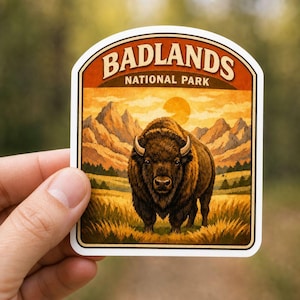 May include: A die-cut sticker featuring a brown bison standing in a grassy field with mountains in the background under a sunset sky. The top of the sticker has an orange banner with the text BADLANDS NATIONAL PARK in white capital letters.