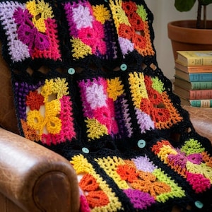 May include: A colorful, handmade crochet blanket with a patchwork design. The blanket features vibrant squares in shades of pink, yellow, orange, and purple, bordered by black crochet. Light blue buttons secure the blanket. The blanket is draped over a leather chair.