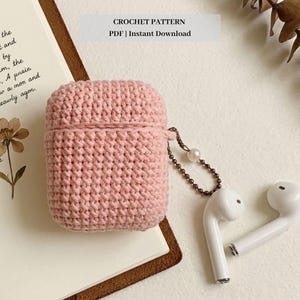 Puede incluir: Una funda para AirPod de ganchillo rosa claro con una tapa a juego. La funda está junto a un par de auriculares inalámbricos blancos y una pequeña cadena con una perla. El texto "CROCHET PATTERN PDF | Instant Download" está en la parte superior.