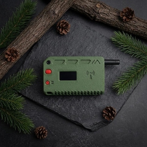 May include: An olive green electronic device with a small screen and red buttons. The device has a black antenna and a wireless signal symbol. The image is styled with pine branches, pine cones, and a dark background.