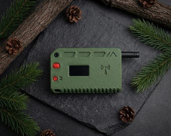 Puede incluir: Un dispositivo electrónico verde oliva con una pequeña pantalla y botones rojos. El dispositivo tiene una antena negra y un símbolo de señal inalámbrica. La imagen está estilizada con ramas de pino, piñas y un fondo oscuro.