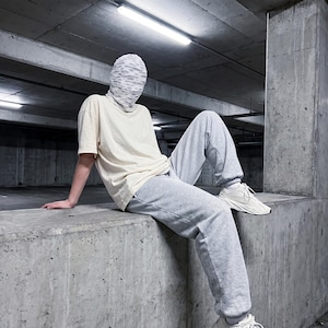 May include: A person wearing a cream-colored t-shirt, gray sweatpants, and white sneakers. They are also wearing a white, textured face covering. The person is sitting on a concrete ledge in a parking garage.