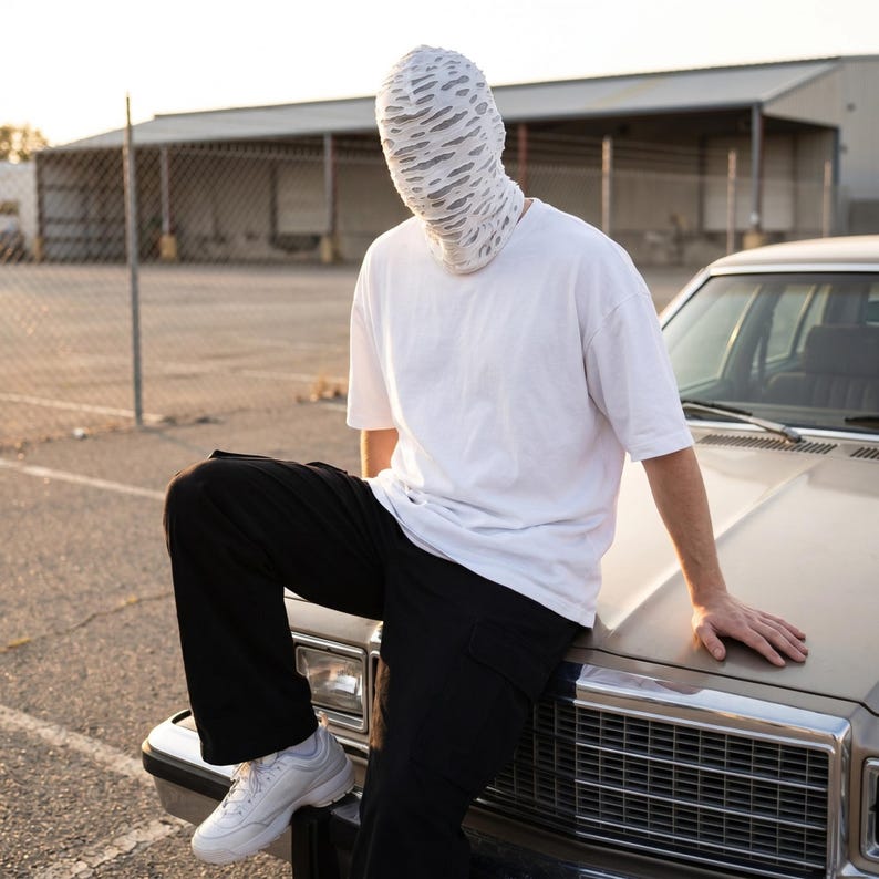 May include: A person wearing a white t-shirt, black pants, and white sneakers, with a textured white head covering. The person is leaning on the hood of a vintage car in an outdoor setting.