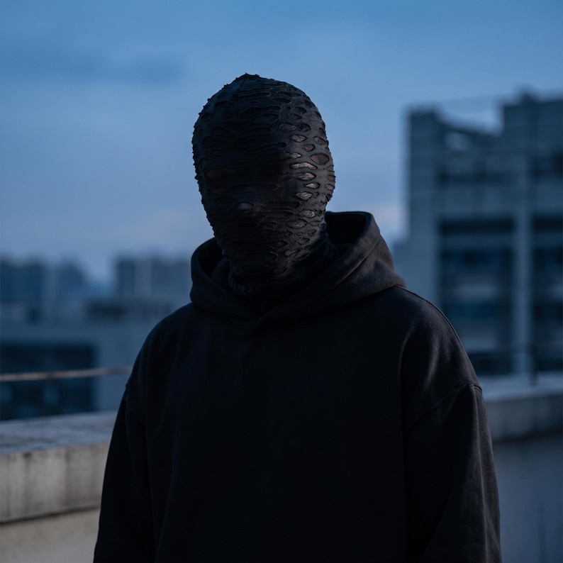 May include: A person wearing a black hoodie and a textured, dark mask. The mask covers the entire head and face, with small openings for the eyes. The background features a cityscape under a dusky sky.