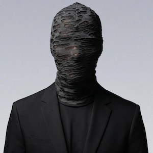 May include: A person wearing a black suit and a black, textured face covering. The face covering obscures the eyes and mouth, creating a mysterious and avant-garde look. The suit has a classic two-button design.