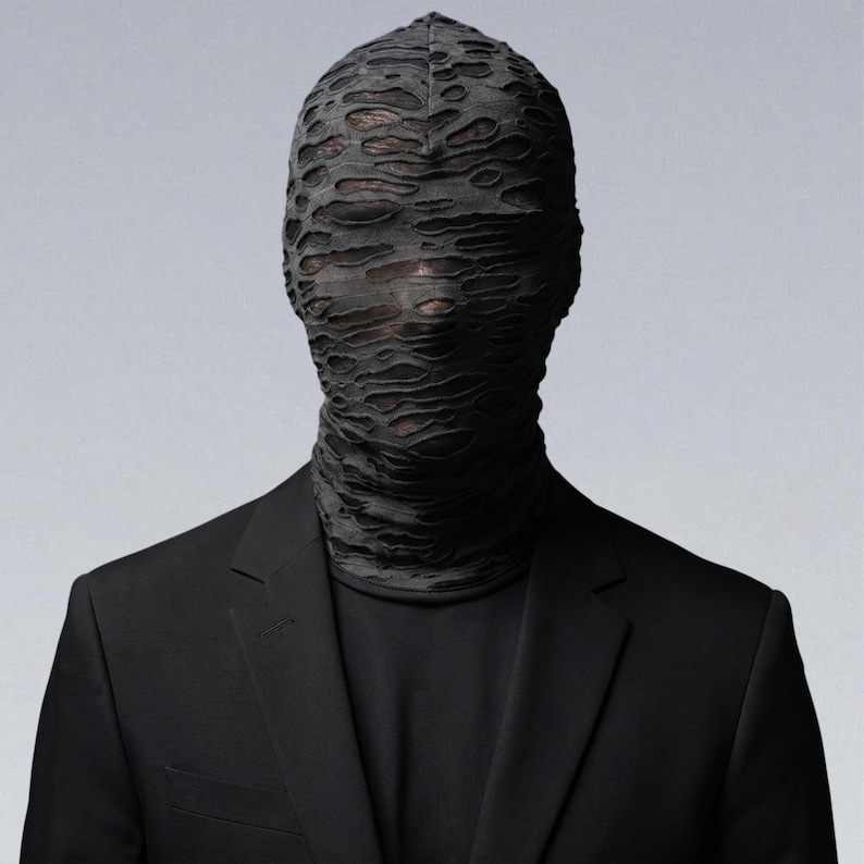 May include: A person wearing a black suit and a black, textured face covering. The face covering obscures the eyes and mouth, creating a mysterious and avant-garde look. The suit has a classic two-button design.