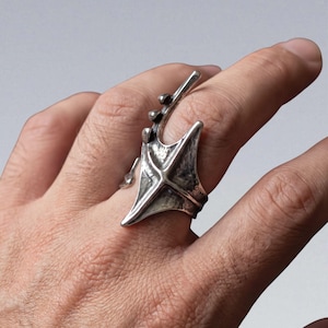 Puede incluir: Un anillo plateado con un diseño asimétrico único. El anillo presenta una forma central estilizada en forma de ala con una superficie texturizada y un elemento lineal elevado que se extiende hacia arriba. El anillo se lleva en un dedo.