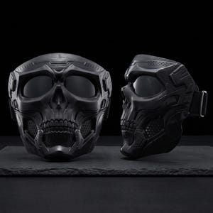May include: Two black skull-shaped masks with dark lenses and intricate detailing. One mask is positioned front-facing, while the other is in profile, showcasing the side strap. The masks are displayed on a dark surface against a black backdrop.
