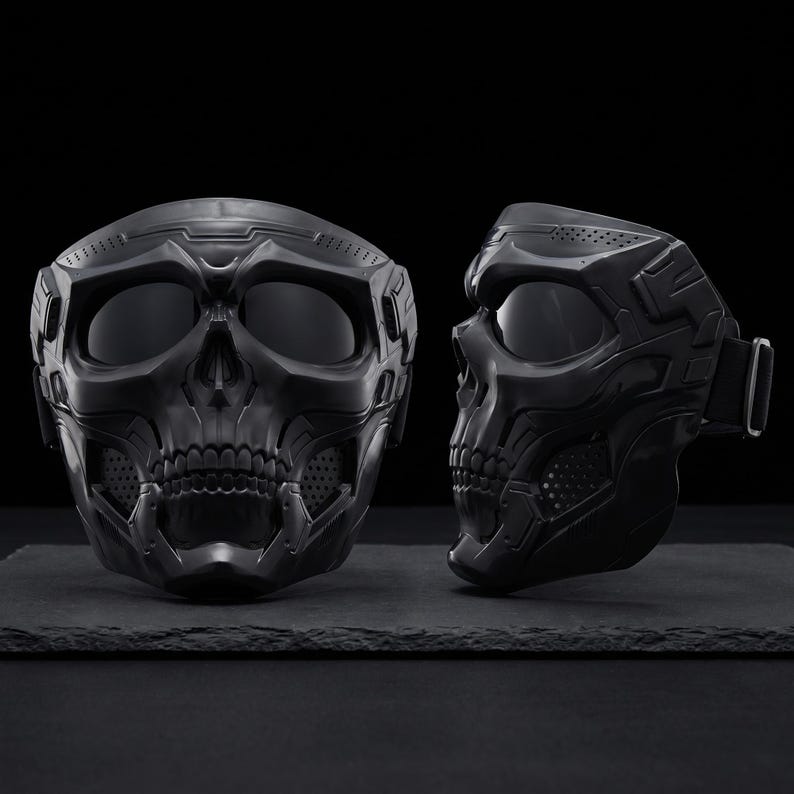 May include: Two black skull-shaped masks with dark lenses and intricate detailing. One mask is positioned front-facing, while the other is in profile, showcasing the side strap. The masks are displayed on a dark surface against a black backdrop.