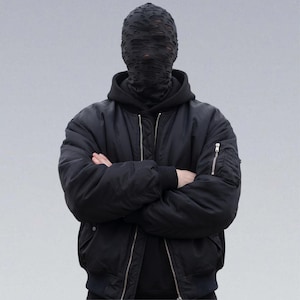 May include: A person wearing a black bomber jacket with a black hooded sweatshirt underneath. The person is also wearing a black face covering with a distressed, torn design. The person's arms are crossed in front of them.