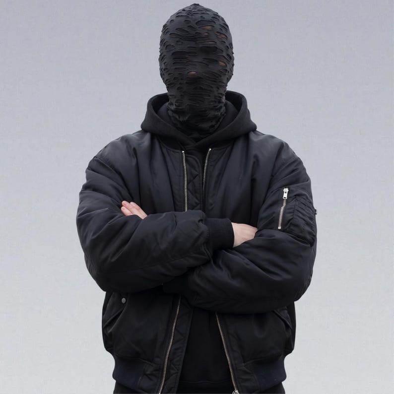 May include: A person wearing a black bomber jacket with a black hooded sweatshirt underneath. The person is also wearing a black face covering with a distressed, torn design. The person's arms are crossed in front of them.
